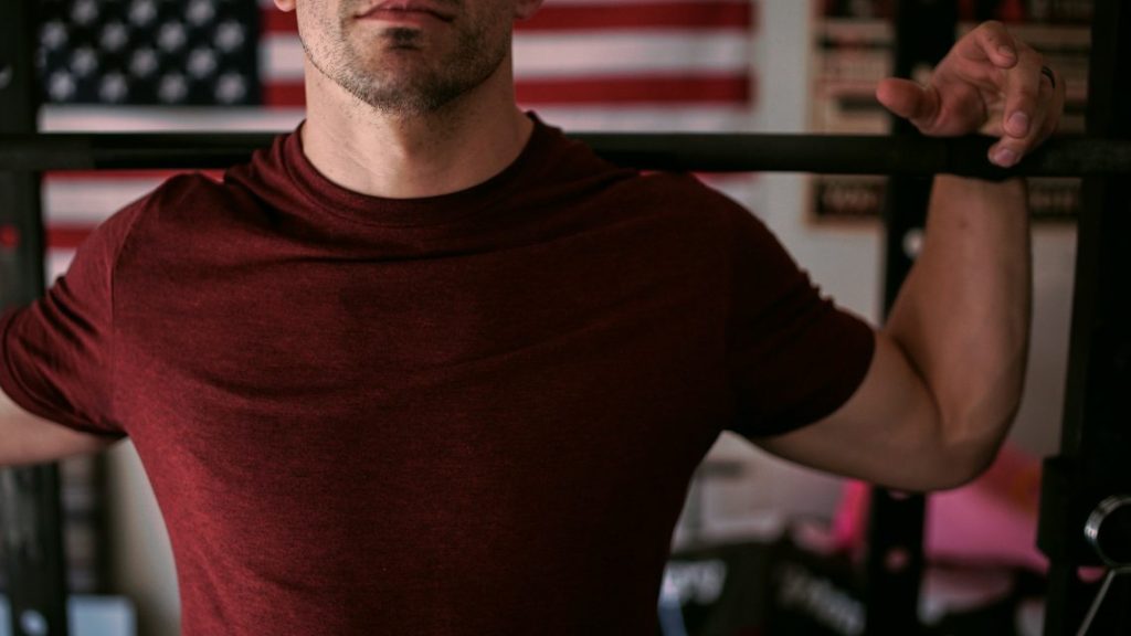 Close-up of man's chest as he holds a barbell behind his shoulders.